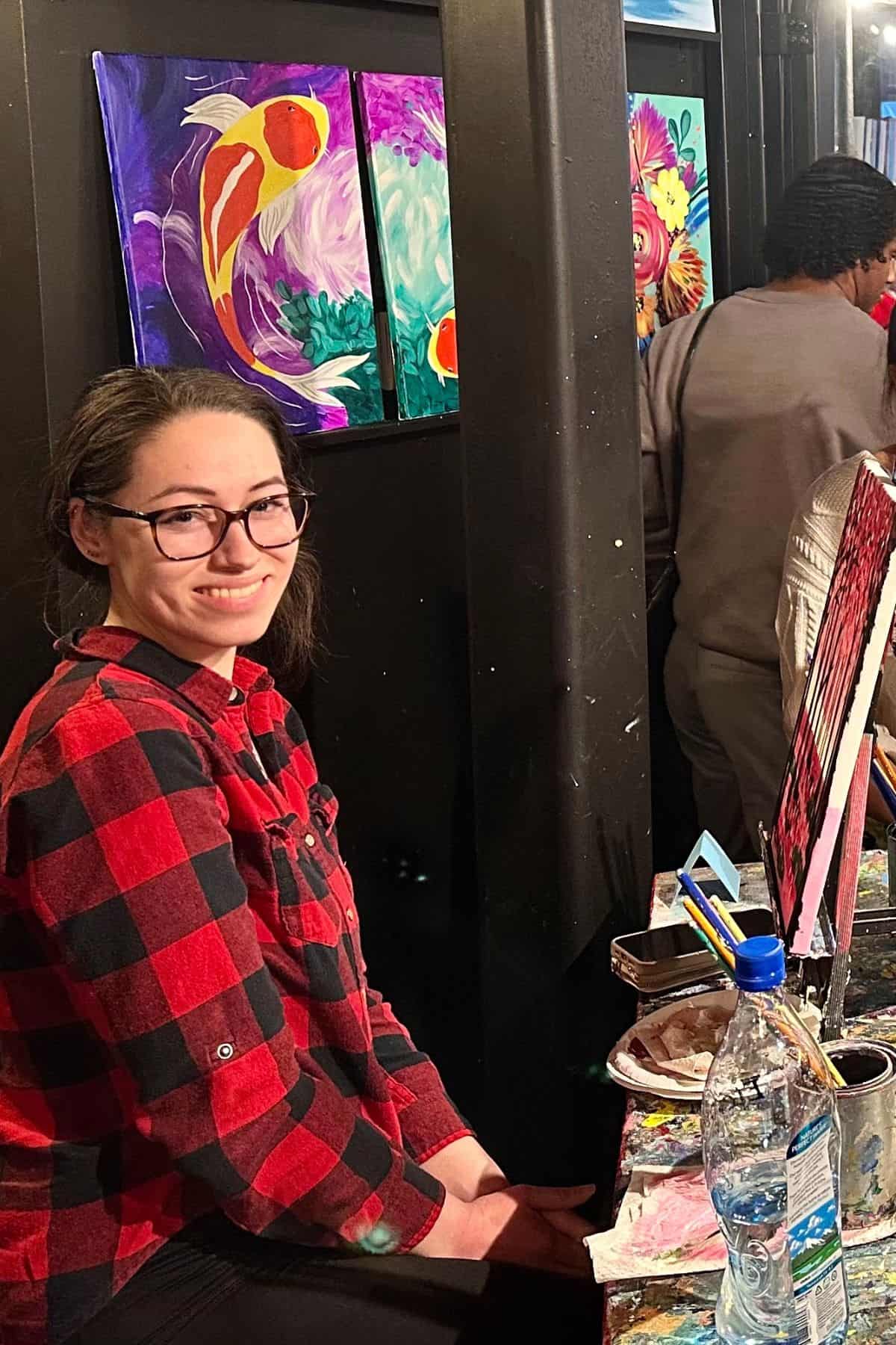 Person in a red and black plaid shirt sits at an artist's station, smiling. Paintings hang on the wall in the background.