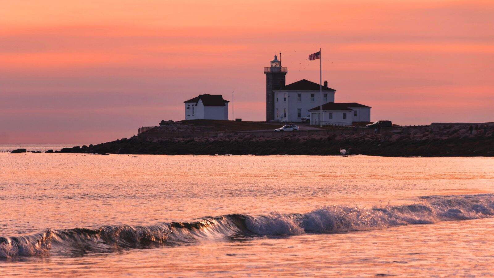 A lighthouse and several buildings sit on a rocky shoreline at sunset with gentle waves in the foreground and an American flag flying on a pole.