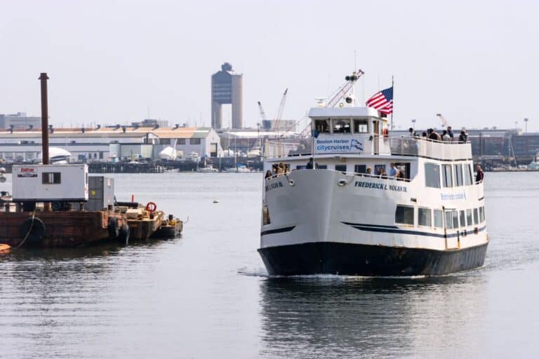A white passenger ferry named Frederick L. Nolan Jr. approaches a dock in an industrial harbor area, with buildings and equipment visible in the background.