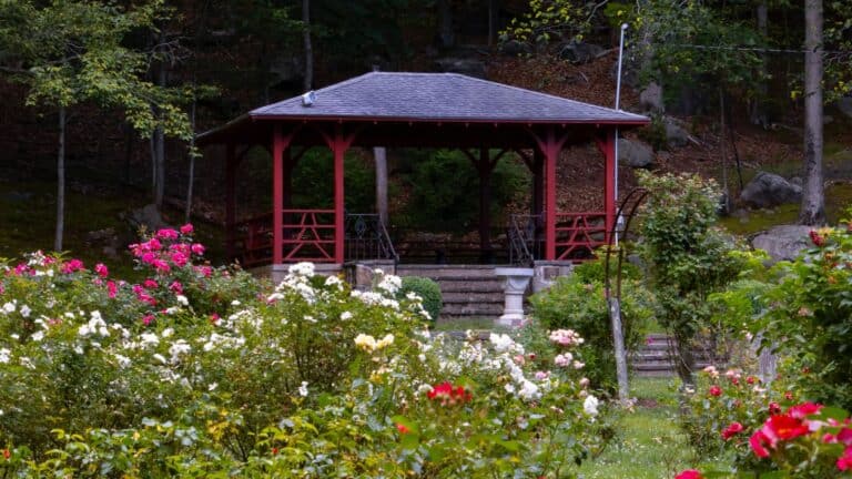 A wooden gazebo stands in a garden filled with red, pink, and white flowers, surrounded by trees and greenery. Steps lead up to the covered structure, which has a sloped roof.