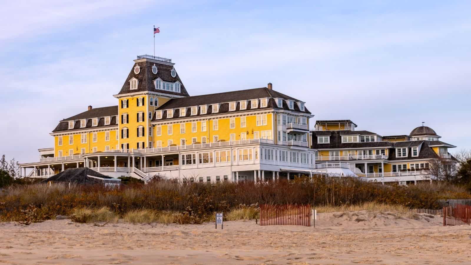 A large yellow and white Victorian-style hotel with an American flag on the roof, situated near sandy dunes and beach vegetation.