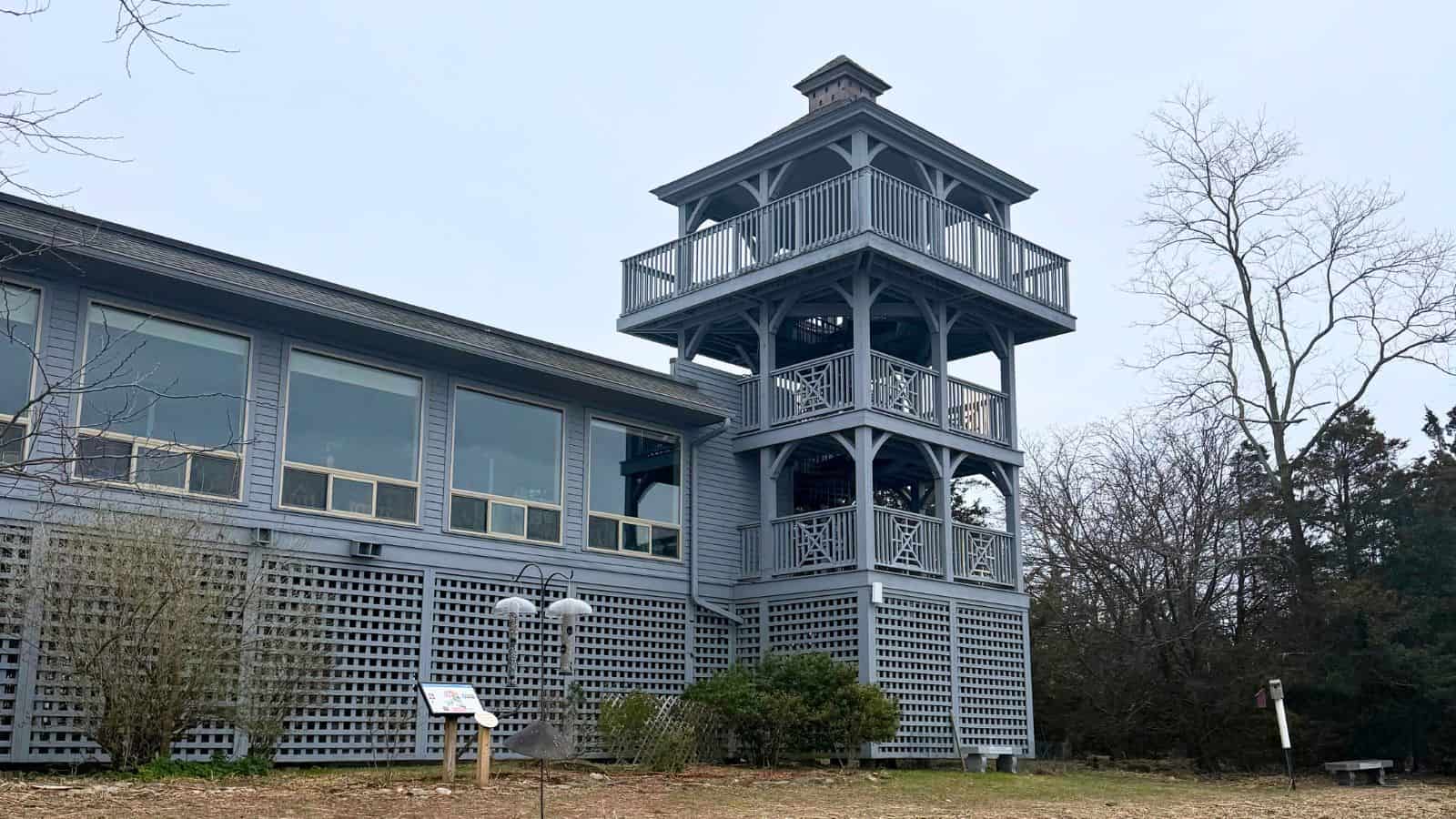 A light blue building with large windows features a tall, multi-level observation tower; trees and shrubs surround the structure on an overcast day.