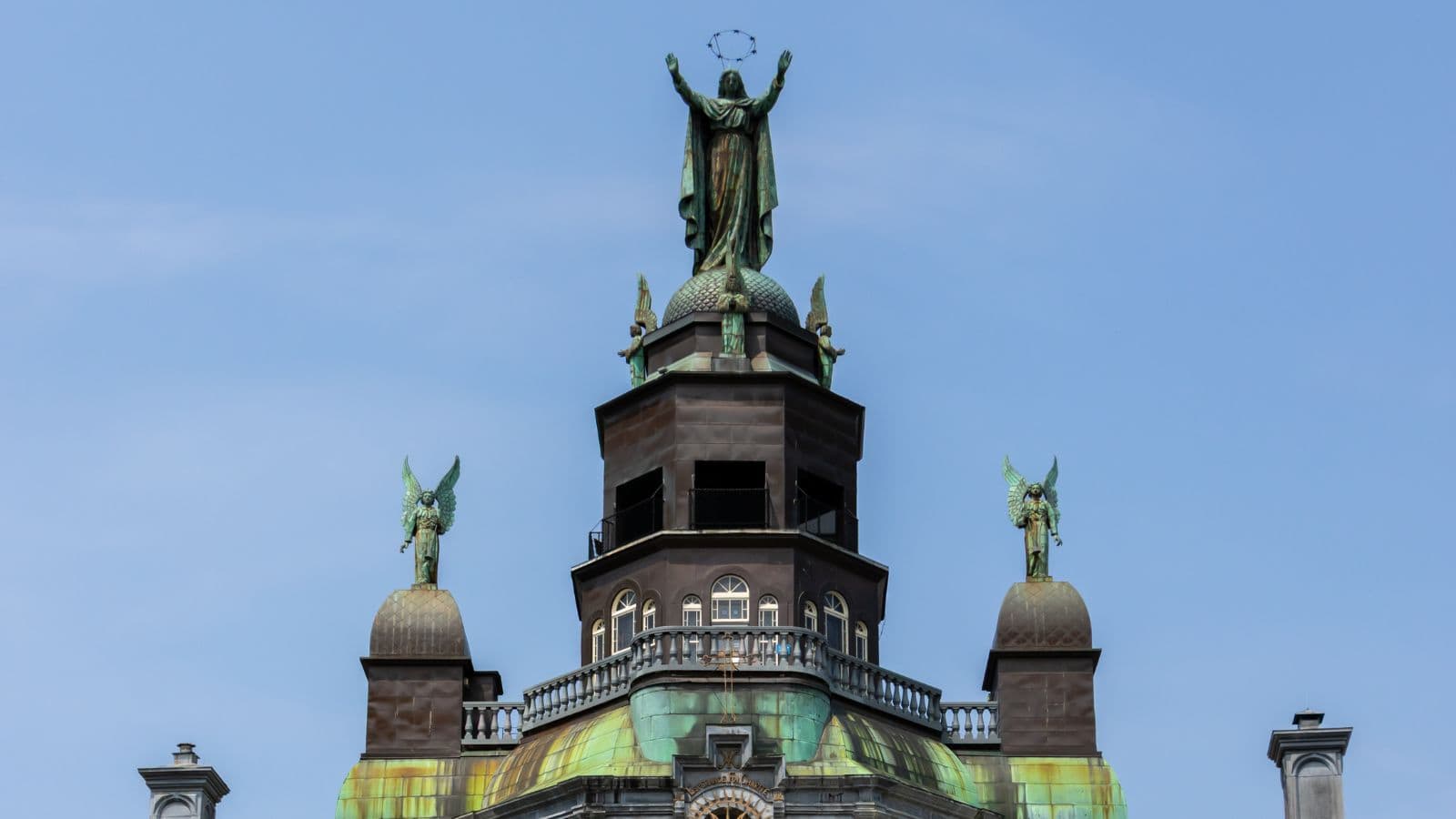 Statue of a robed figure holding a circular object stands atop the historic Notre-Dame-de-Bon-Secours Chapel, with green copper roof accents and two winged statues on either side, set against a clear blue sky.