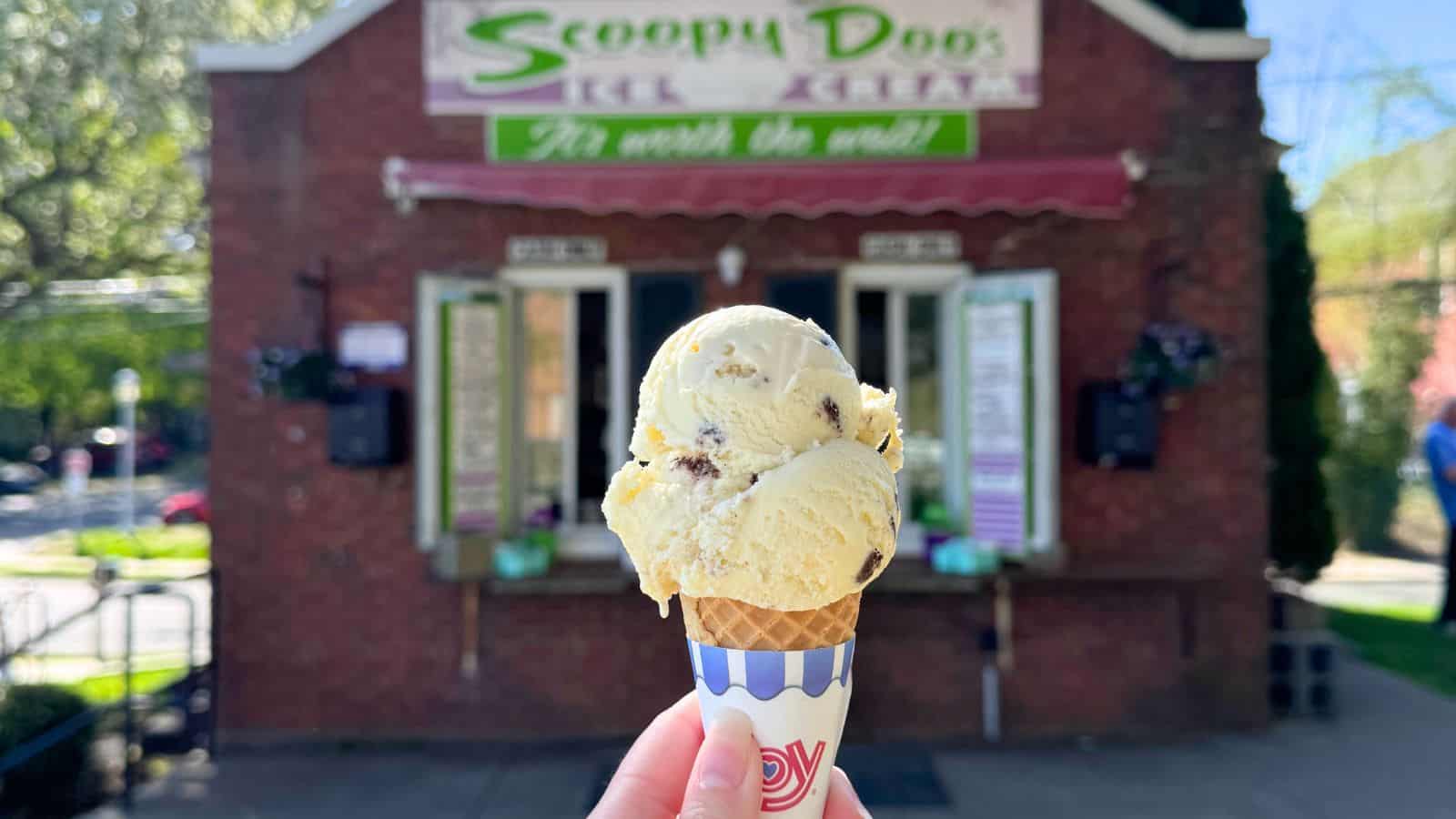 A hand holds a vanilla ice cream cone with chocolate chips in front of a small brick ice cream shop named Scoopy Doo's.