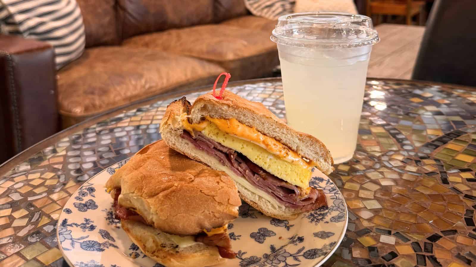 A breakfast sandwich with ham, egg, and cheese is cut in half on a floral plate, next to a plastic cup of lemonade on a mosaic table.