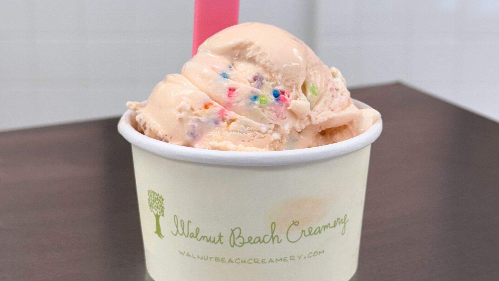 A cup of peach-colored ice cream with colorful sprinkles in a white paper cup labeled "Walnut Beach Creamery," sitting on a dark surface with a pink spoon.