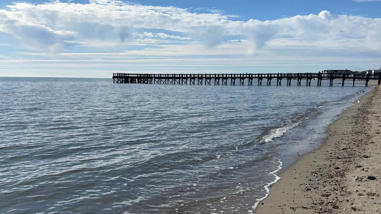 A wooden pier extends over calm water under a partly cloudy sky, with gentle waves reaching a sandy beach in the foreground.
