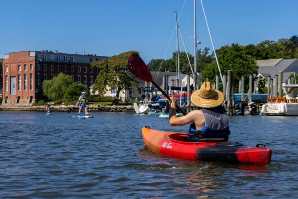A person wearing a straw hat paddles a red kayak on a calm body of water near sailboats, buildings, and people paddleboarding under a clear blue sky.