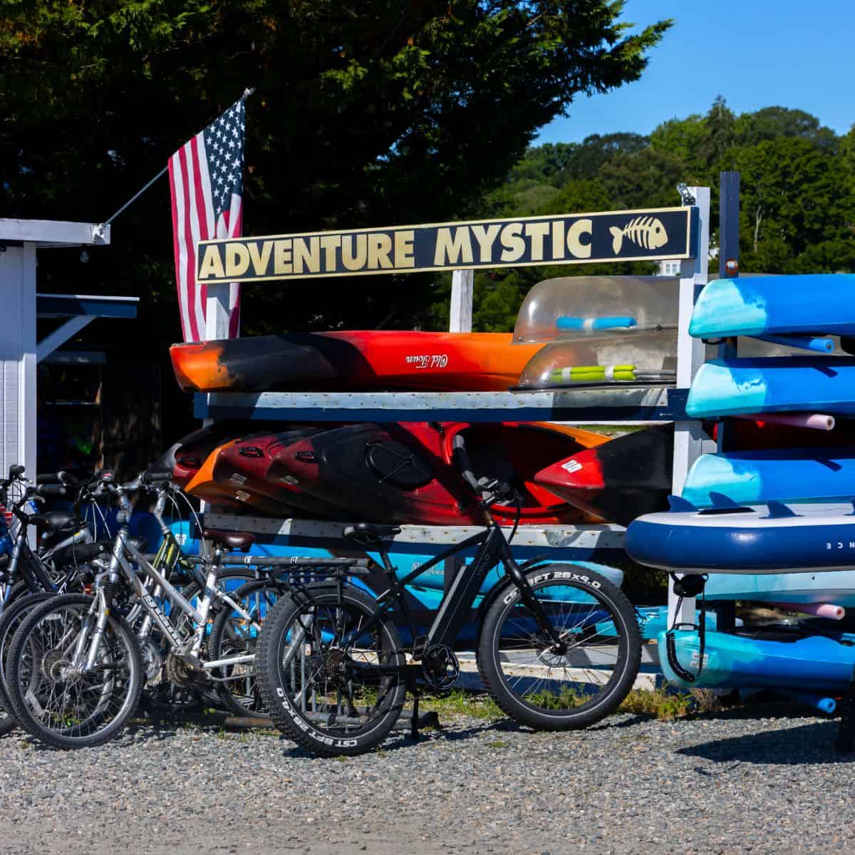 Bicycles and colorful kayaks are displayed outside under a sign reading "Adventure Mystic," with an American flag in the background.