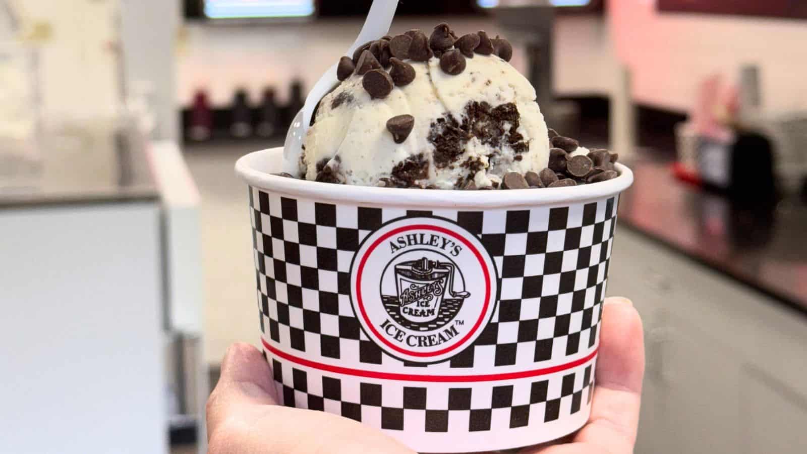 A hand holds a cup of Ashley's Ice Cream topped with chocolate chips and cookie crumbles, with a white spoon in the ice cream.