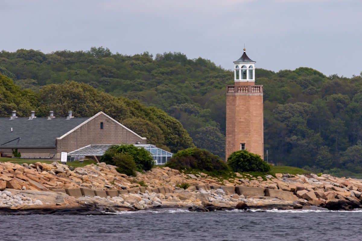 A brick lighthouse with a white lantern room stands on a rocky shoreline near a building, with dense green trees in the background and water in the foreground.