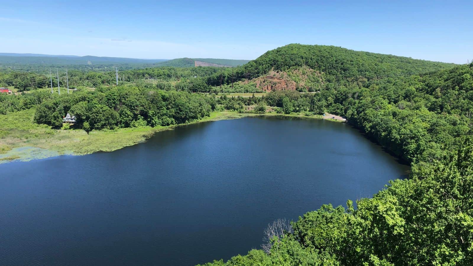 A round, tree-lined lake is surrounded by green hills and forest under a clear blue sky on a sunny day.