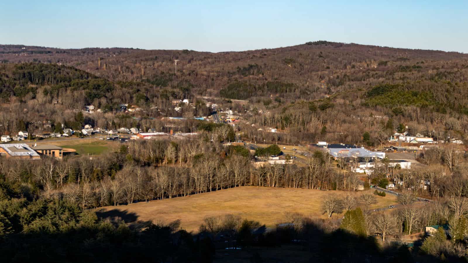 Aerial view of a small rural town with open fields, scattered houses, and tree-covered hills under a clear sky.