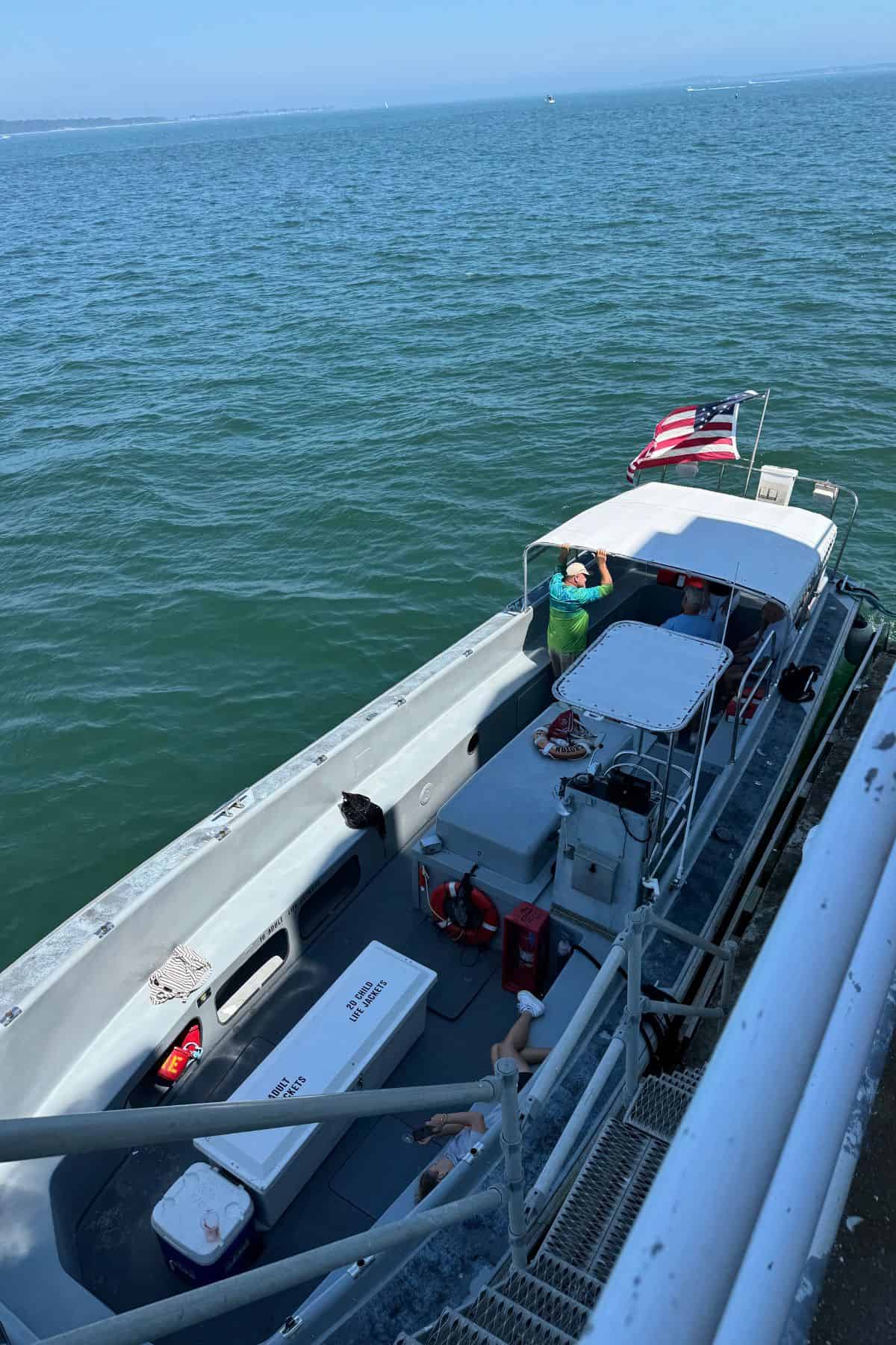 A small motorboat with a U.S. flag is docked beside a metal staircase; a person in a green shirt stands near the helm, and coolers are visible on the deck. The boat is on calm water.