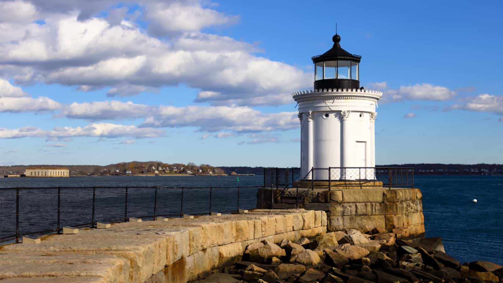 A white lighthouse stands at the end of a stone pier overlooking a calm, blue body of water under a partly cloudy sky.