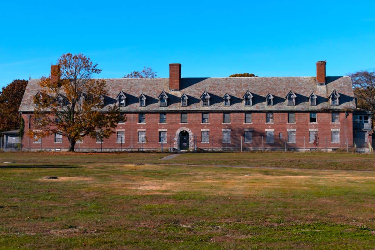A large, old red-brick building with boarded windows and chimneys, set behind a fenced grassy field under a clear blue sky.
