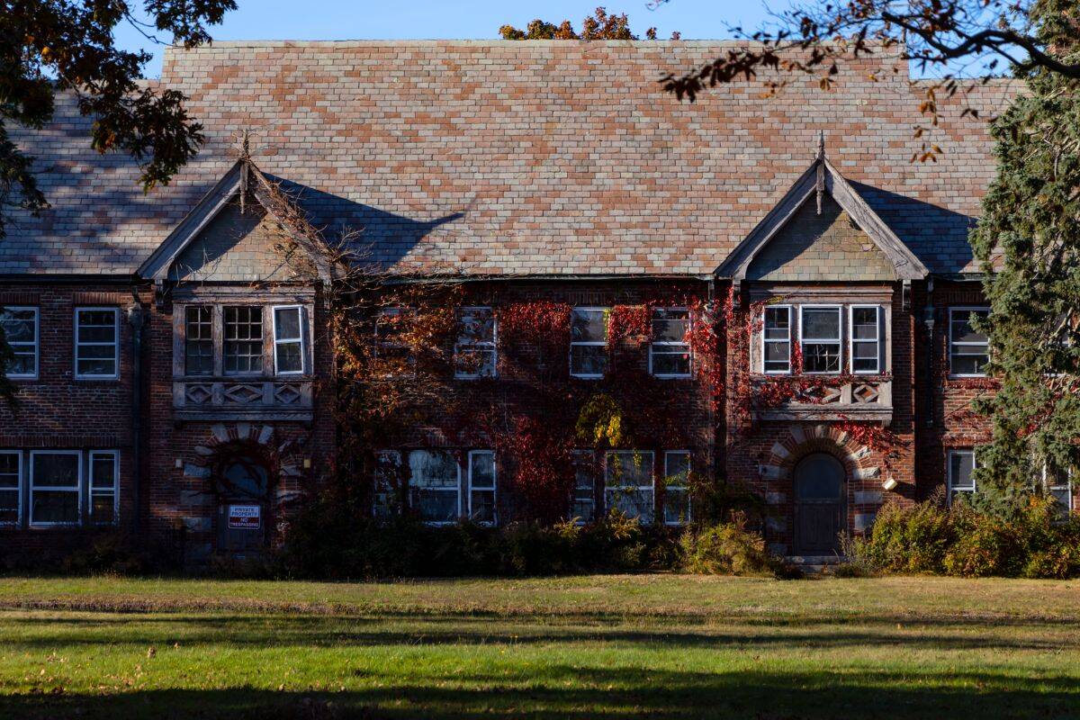 Old brick building with a shingled roof, arched doorways, and windows; some ivy grows on the facade. Grass lawn and shadows are visible in the foreground.