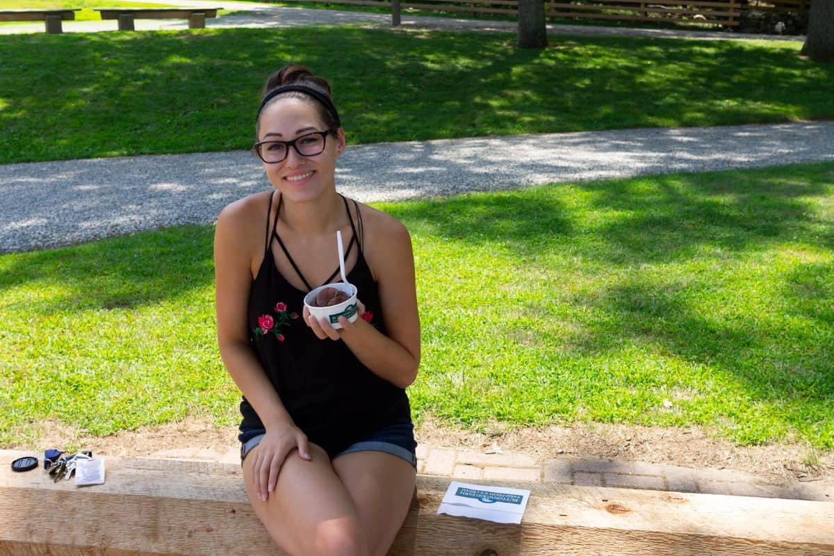 A woman wearing glasses and a black tank top sits on a wooden bench outside, holding a cup of food with a spoon. Keys and a paper are on the bench beside her.