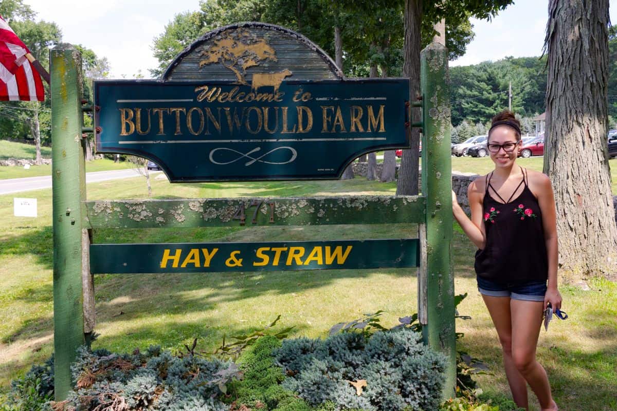 A woman stands next to a green sign that reads "Welcome to Buttonwood Farm" and "Hay & Straw," with trees and grass in the background.