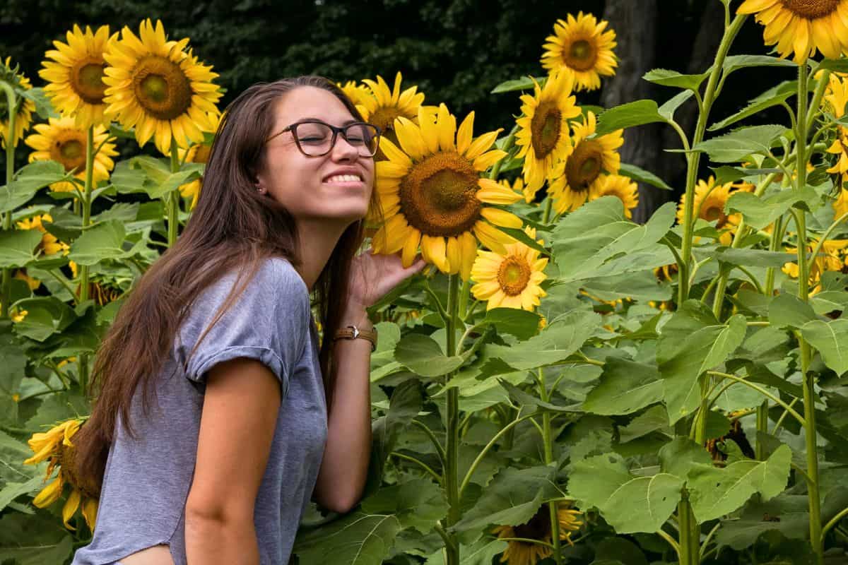 A woman with long brown hair and glasses smiles while posing next to a sunflower in a field of sunflowers.