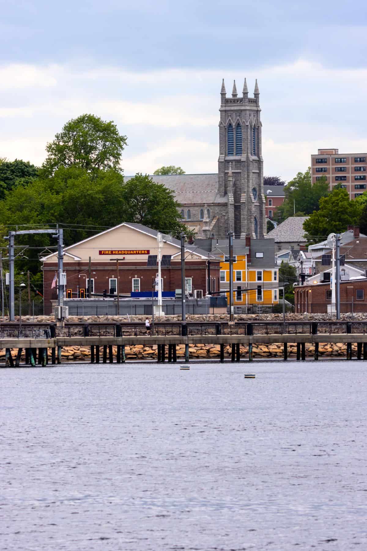 A waterfront scene with a pier, buildings, and a tall stone church with a clock tower in the background, surrounded by trees and other structures.