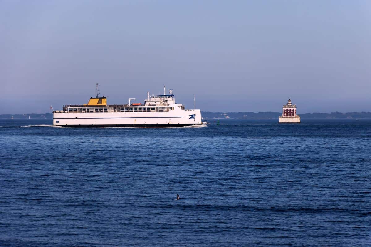 A large white ferry travels across a calm body of water near a small lighthouse structure, with distant shoreline visible under a clear sky.