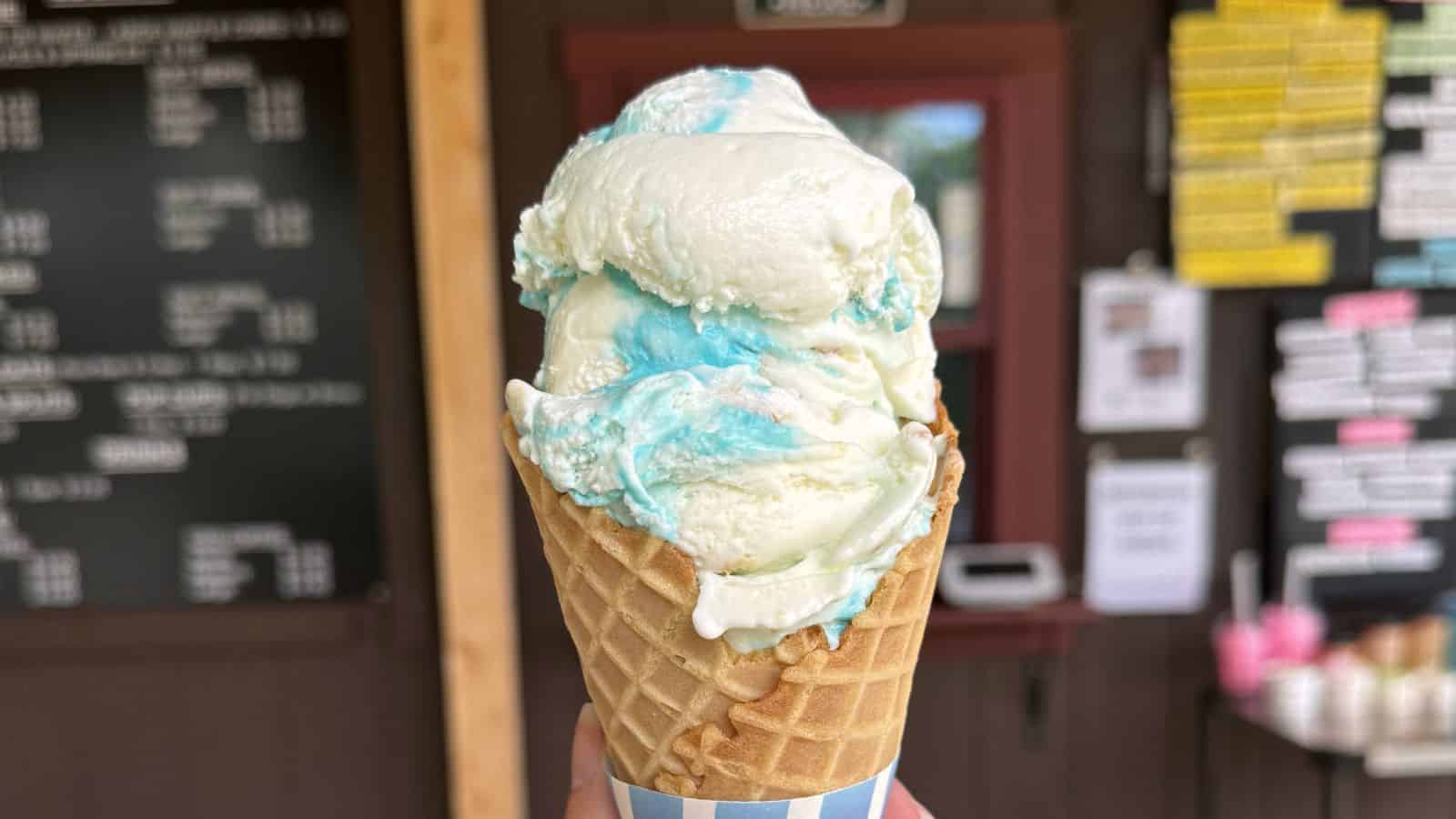 A close-up of a waffle cone with two scoops of white and blue swirled ice cream, held up in front of a menu board at an ice cream shop.