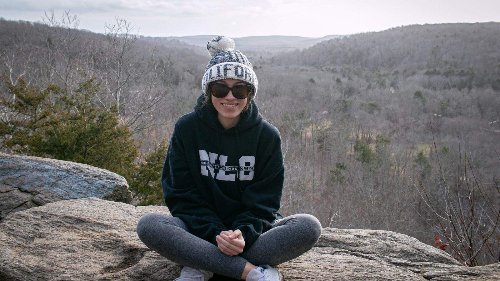 A person wearing a beanie, sunglasses, and hoodie sits cross-legged on a rock with a forested landscape in the background.