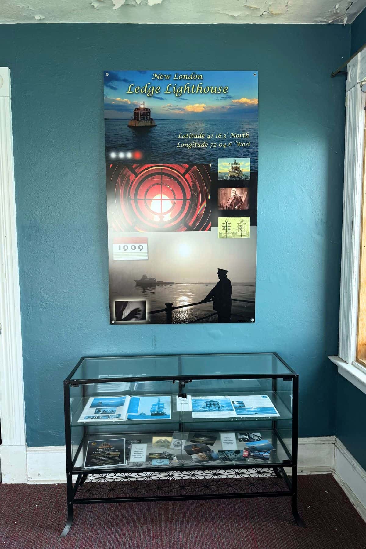 Glass display case with brochures and artifacts beneath a wall-mounted informational poster about the New London Ledge Lighthouse, featuring images and details about the lighthouse.