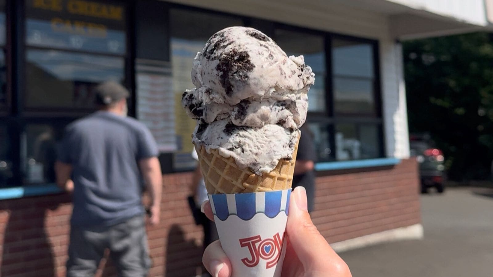 A hand holds up a cookies and cream ice cream cone in front of an ice cream shop, one of the best ice cream spots in Connecticut, with two people standing in line.
