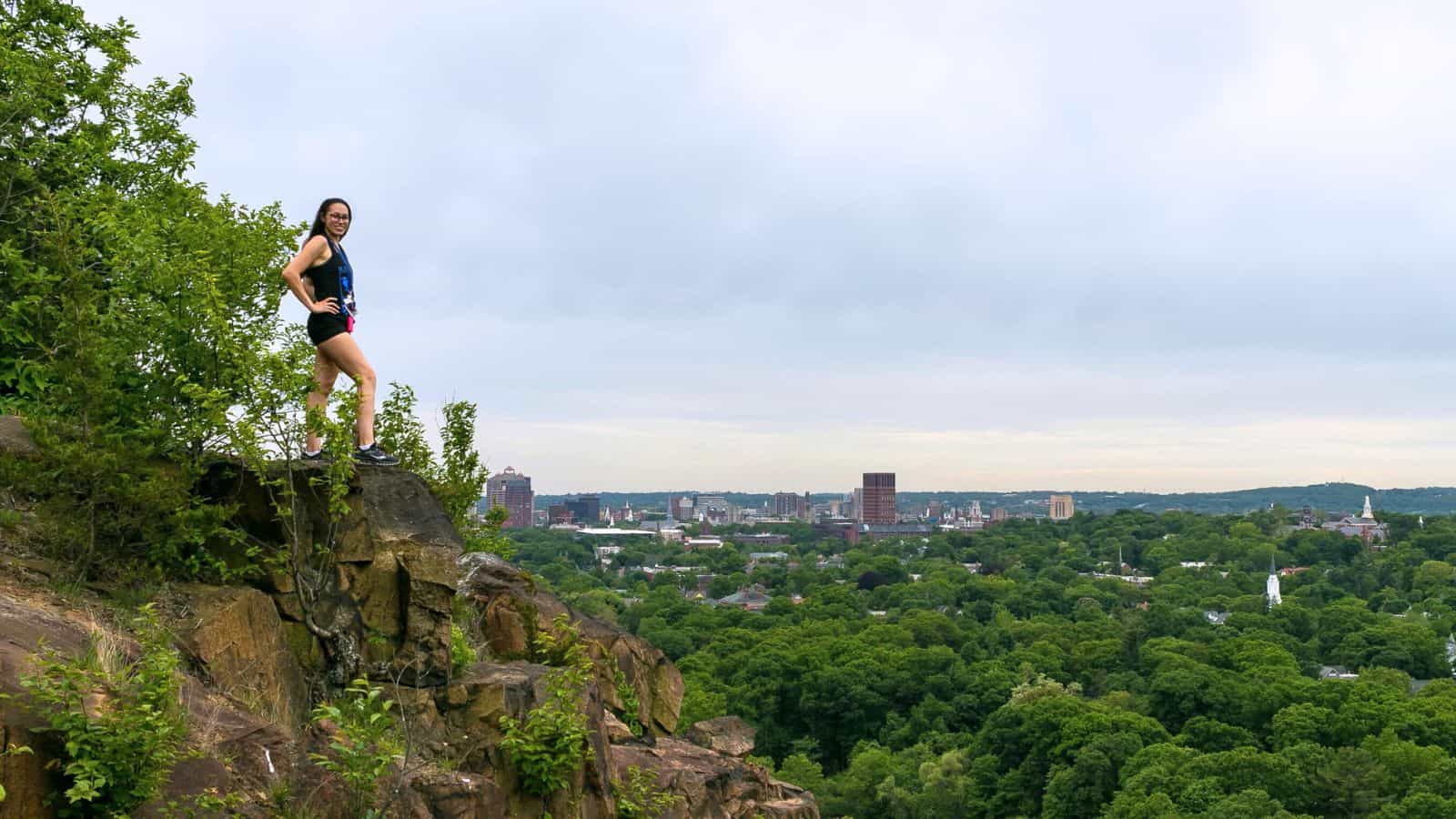 A person stands on a rocky cliff edge overlooking a city skyline and dense green forest under a cloudy sky.