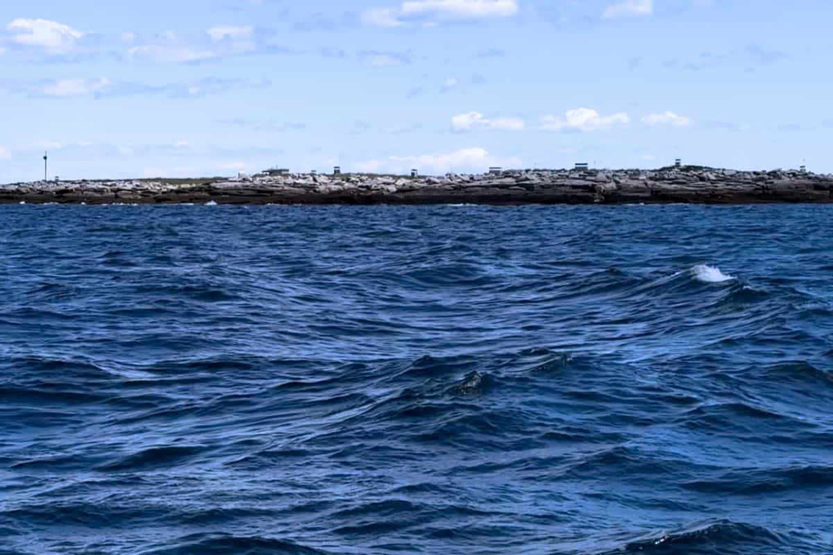 Choppy blue water in the foreground with a rocky, low-lying shoreline and a partly cloudy sky in the background.