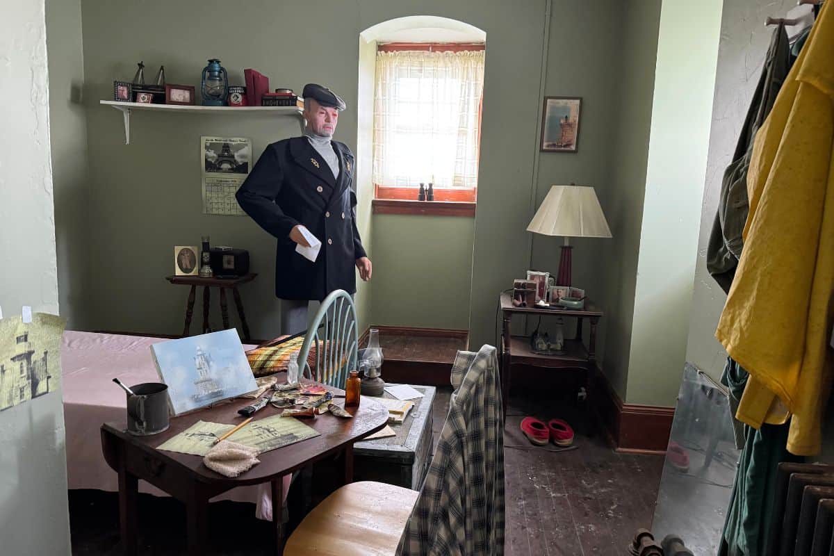 A mannequin dressed in a vintage military uniform stands in a small, cluttered room with a single bed, table, chair, and various personal items.