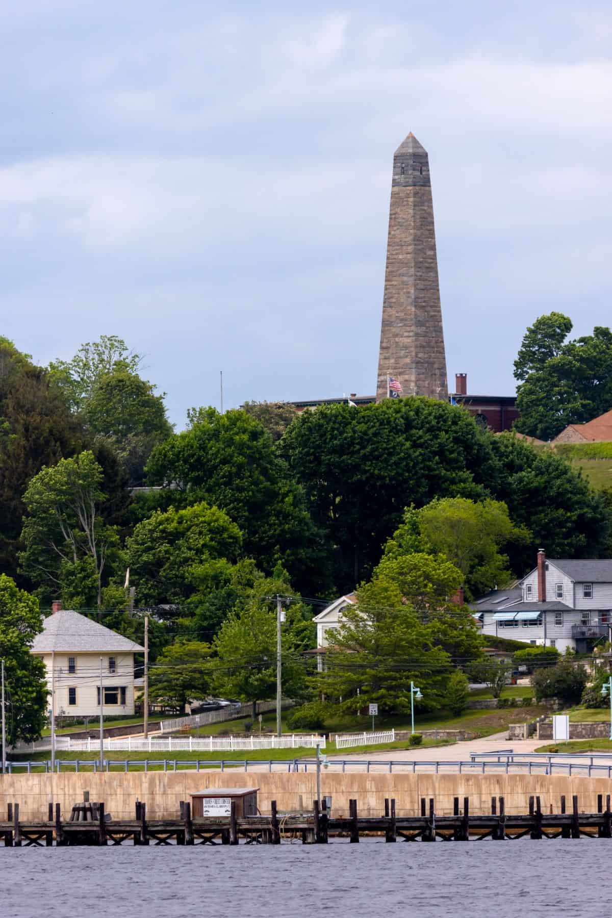 Tall stone obelisk monument rises above a shoreline with houses, trees, and a waterfront dock under a cloudy sky.