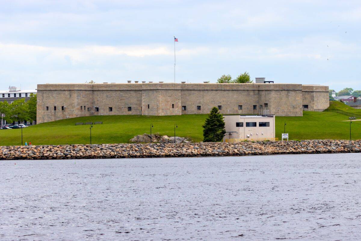 A stone fort with an American flag on top, surrounded by grass and a stone seawall, sits next to a body of water under a cloudy sky.