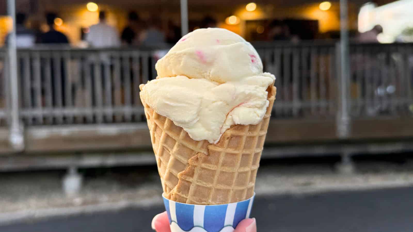A hand holds a waffle ice cream cone with two scoops of vanilla ice cream in front of a blurred outdoor seating area with warm lights.