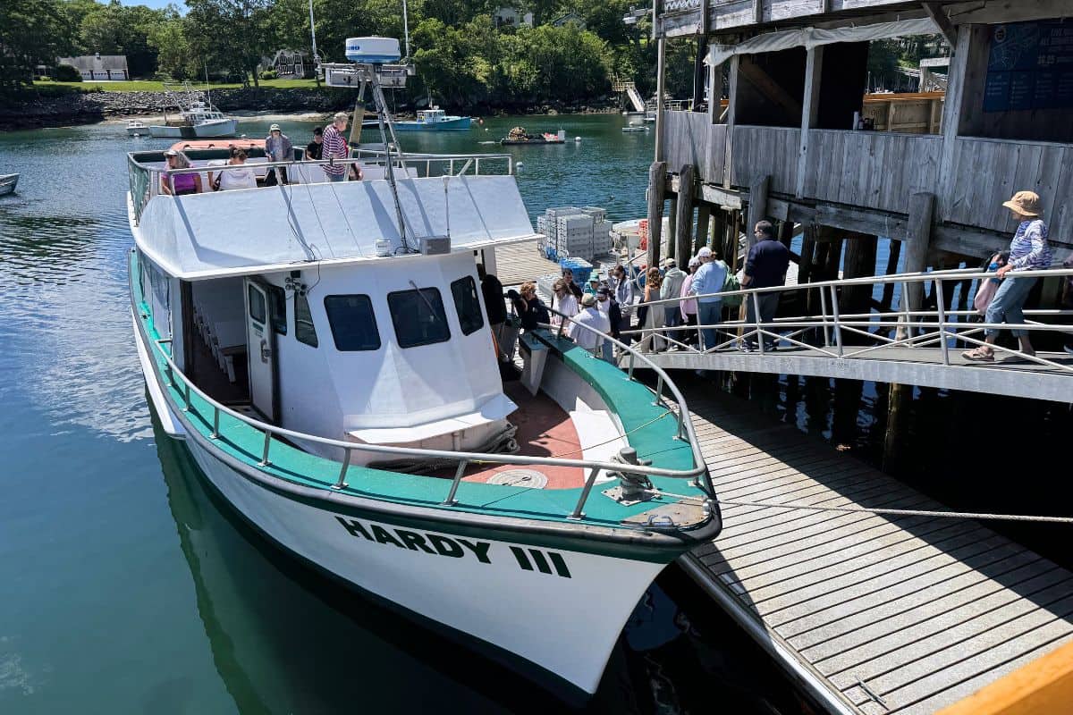 A white and green boat named HARDY III is docked by a wooden pier as people board via a ramp on a sunny day.