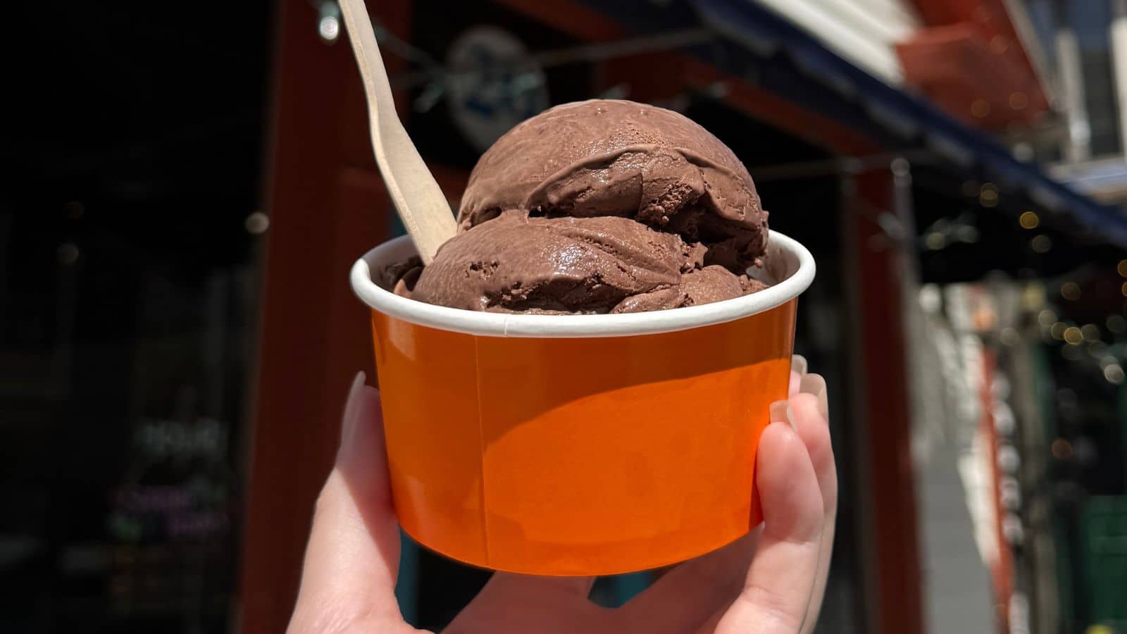 A hand holds an orange cup filled with two scoops of chocolate ice cream and a wooden spoon, with a blurred outdoor background.