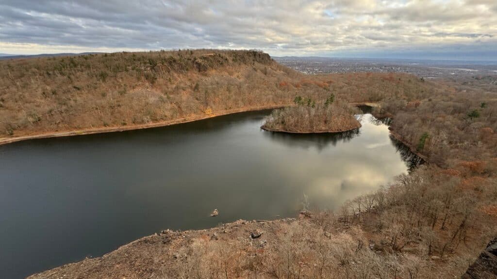 Aerial view of a lake surrounded by bare trees and hills under a mostly cloudy sky, with a small wooded island near the center of the lake.