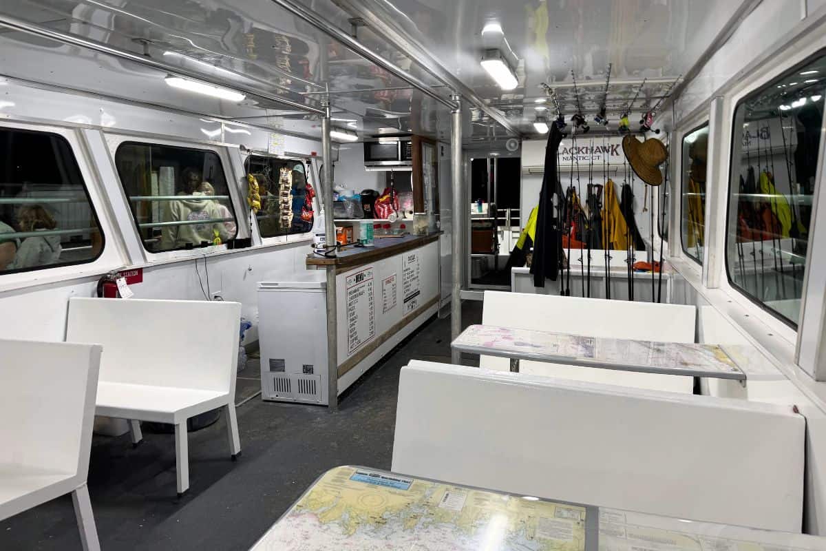 Interior of a boat with white benches and tables, fishing rods lined up on one side, a counter with menus, and equipment hanging on the wall.