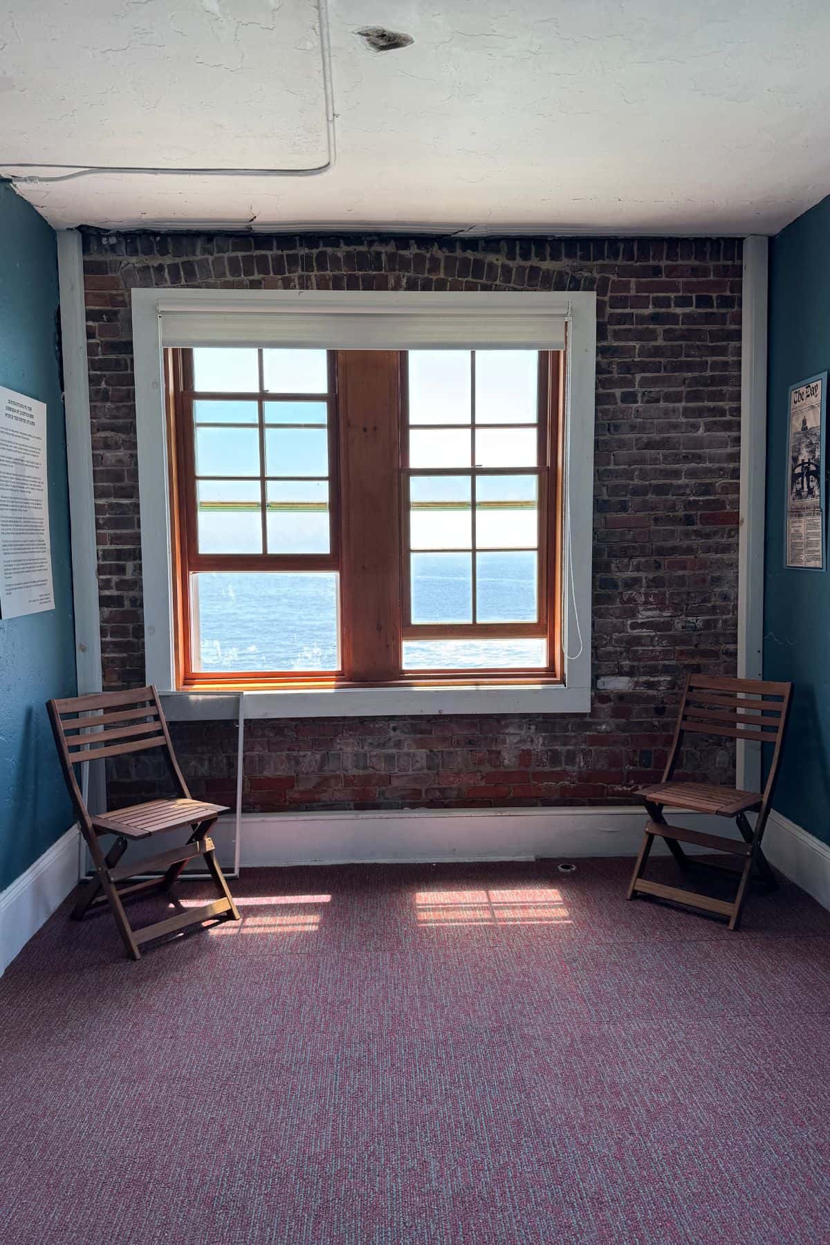 Two wooden chairs face each other in front of a large window with an ocean view, set against an exposed brick wall in a bright, carpeted room.