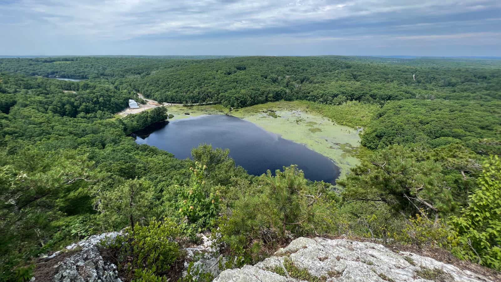 A view from atop a rocky cliff overlooks a small lake surrounded by dense green forest under a partly cloudy sky. A building sits near the lake’s edge.