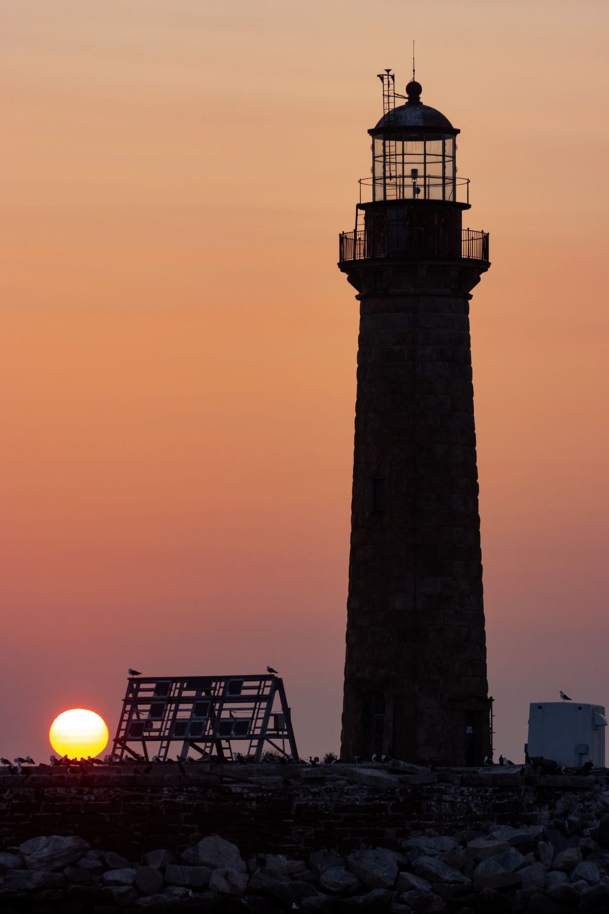 A tall lighthouse stands on a rocky shore at sunset, with the sun low on the horizon and the sky displaying warm orange hues.