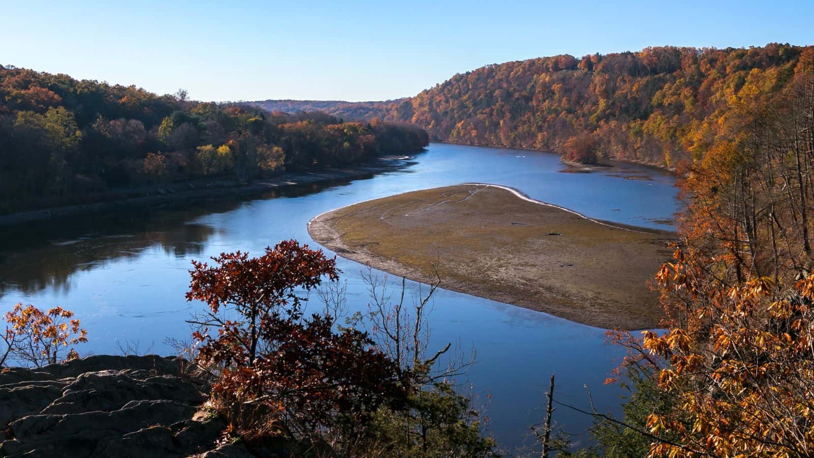 A wide river curves through a forested landscape with autumn-colored trees and a sandy riverbank exposed in the center. The sky is clear and blue.
