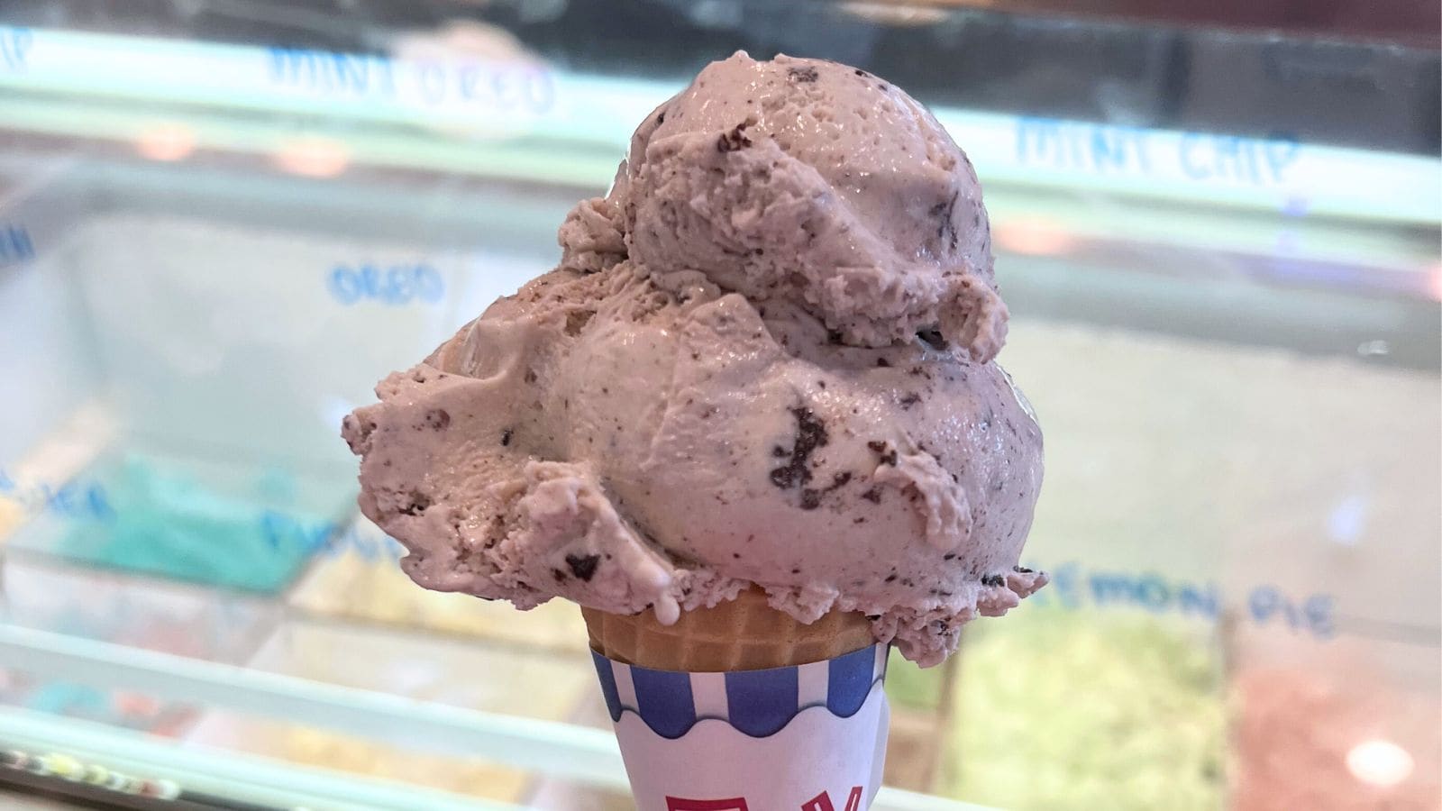 A scoop of cookies and cream ice cream in a waffle cone, held in front of an ice cream display case with various flavors.