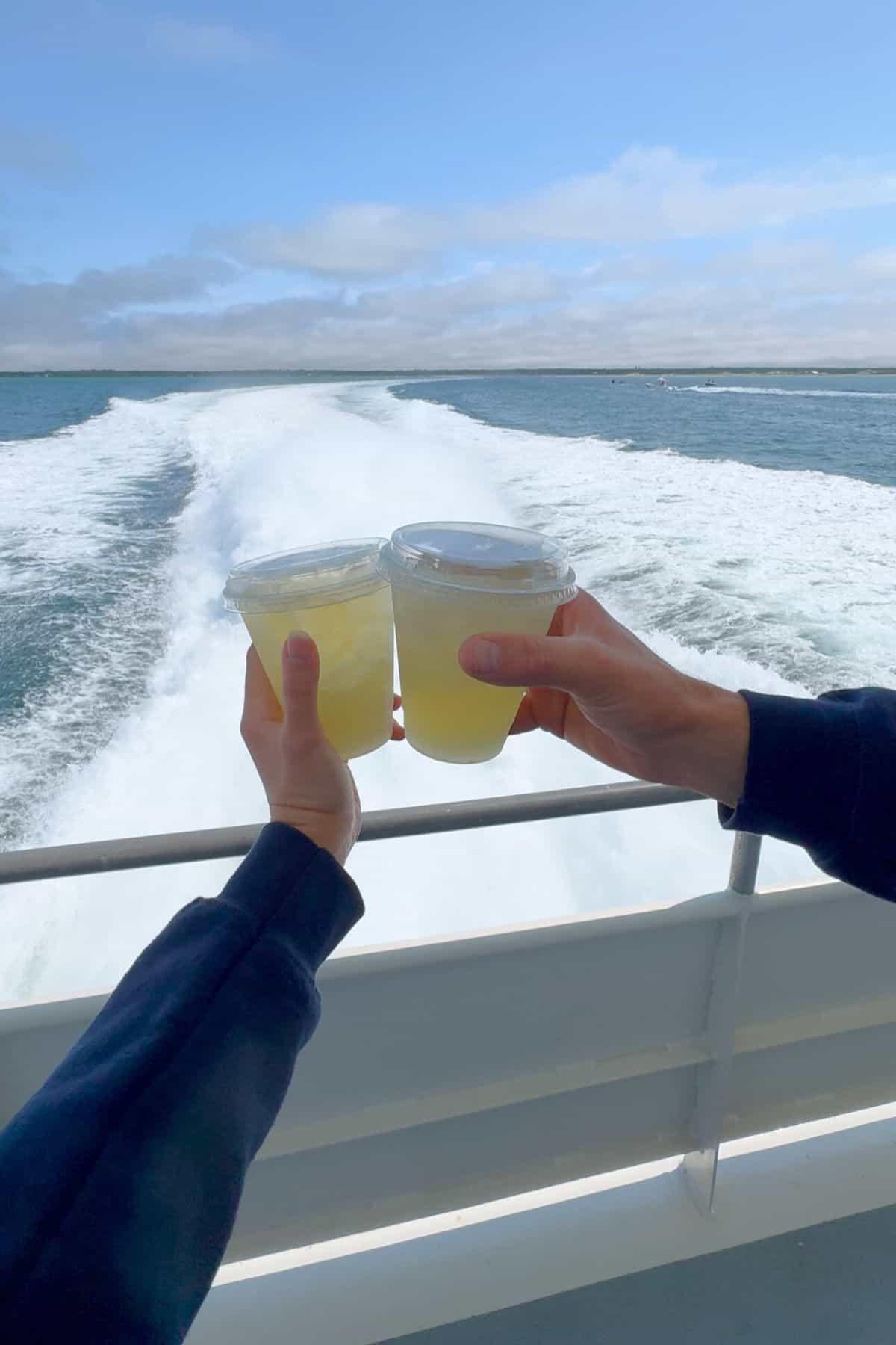 Two people hold up plastic cups of a yellow drink in a toast on a boat, with the ocean and the boat's wake visible in the background under a cloudy sky.
