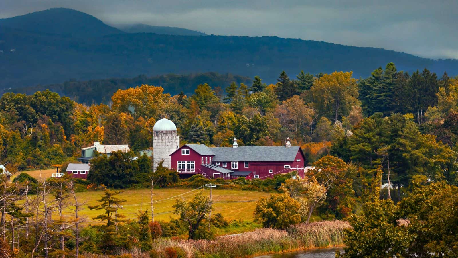 A red barn with a silo sits among trees with autumn foliage, set against a backdrop of forested hills and cloudy skies.