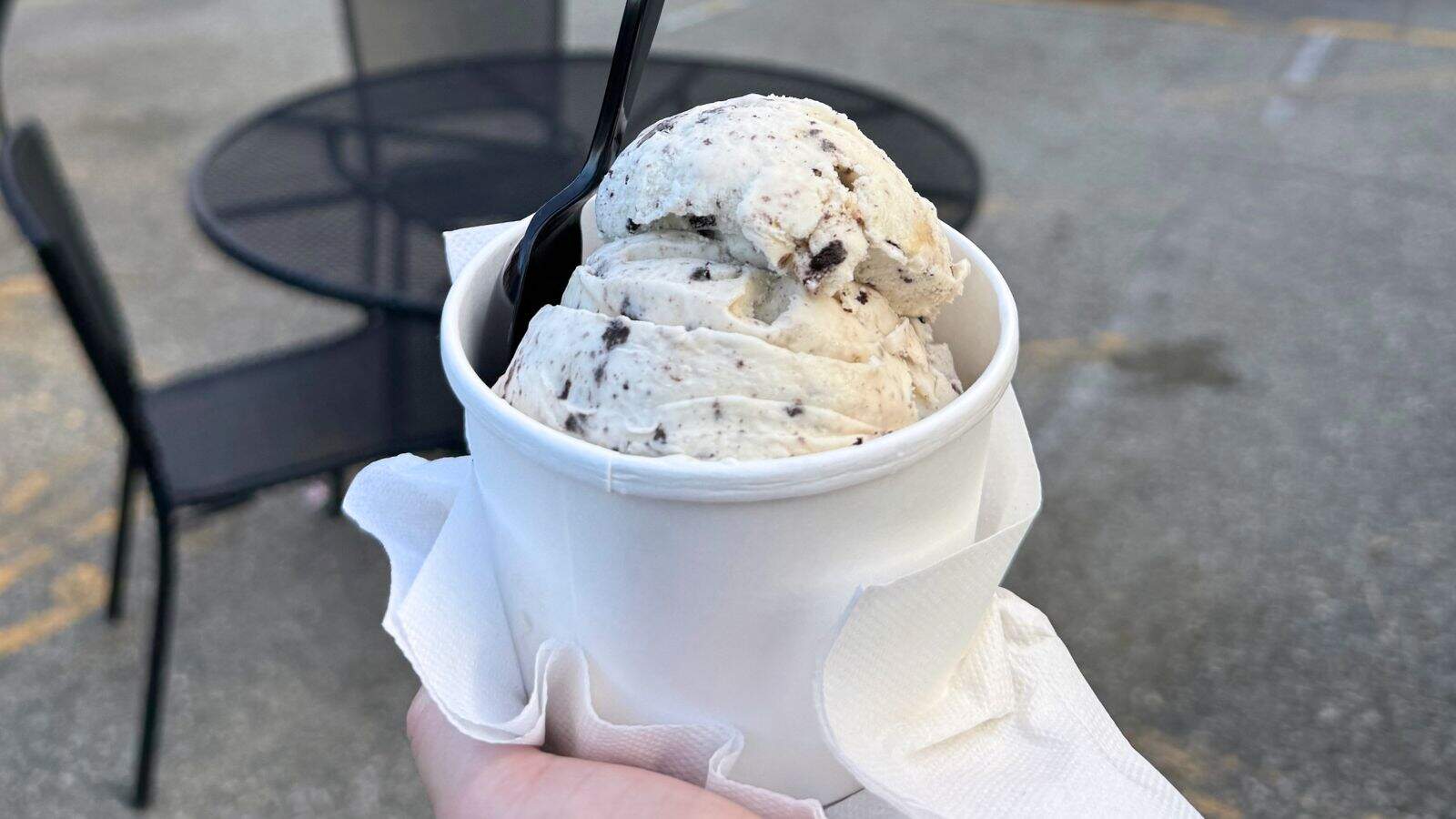 A hand holds a cup of cookies and cream ice cream with a black plastic spoon, next to a napkin, with outdoor tables and chairs in the background.