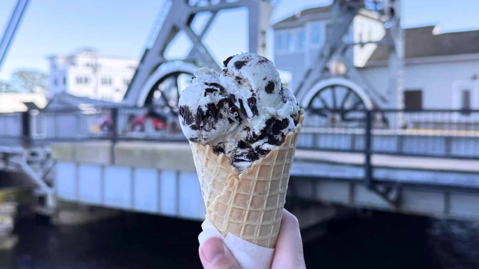 A hand holds a waffle cone with cookies and cream ice cream in front of a metal drawbridge on a sunny day.