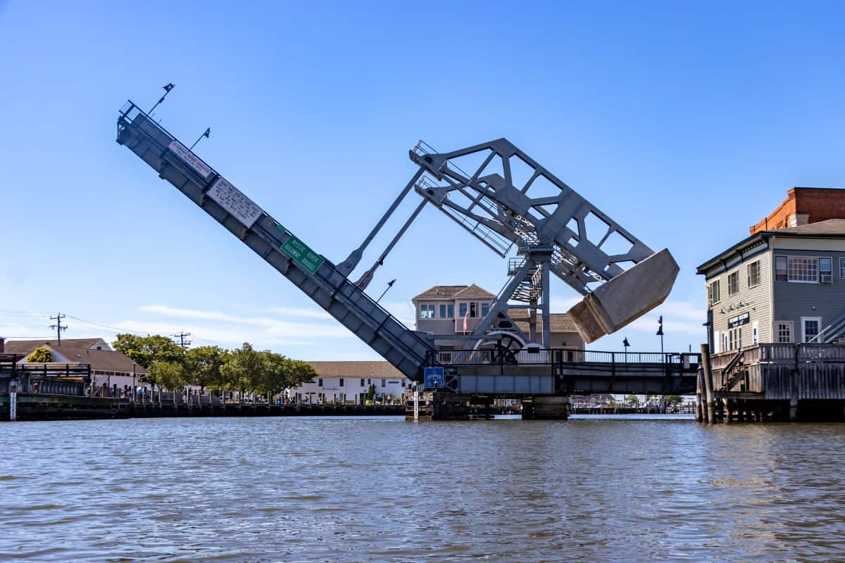 A drawbridge is raised to allow boats to pass underneath on a sunny day, with buildings and trees visible on either side of the water.
